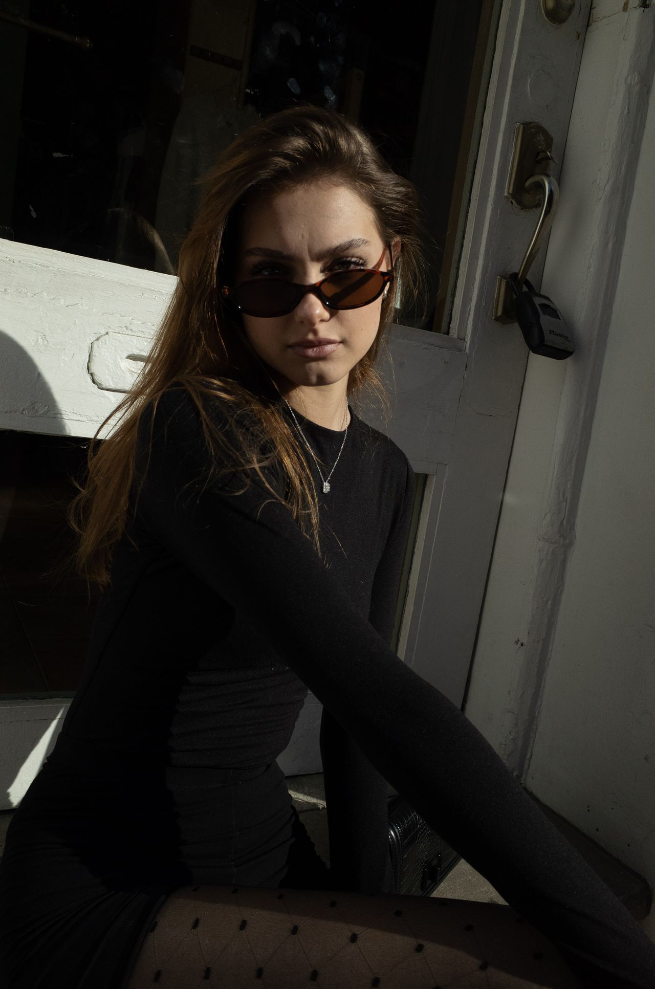 Mia Fantalin seated in a weathered white doorway in hard sunlight, wearing black and tortoise-shell sunglasses