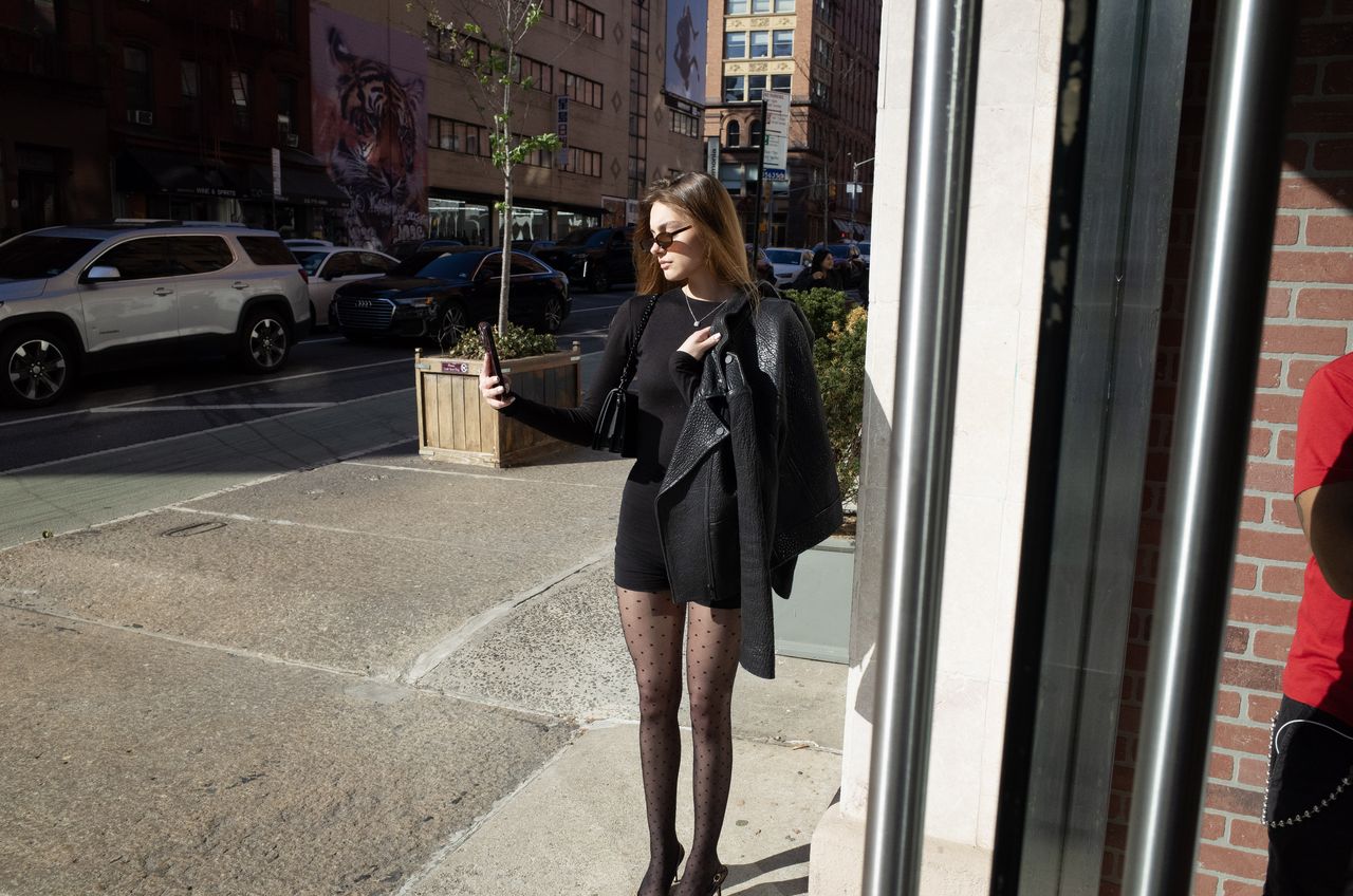 Mia Fantalin standing on a New York sidewalk dressed in black, a leather jacket over one arm