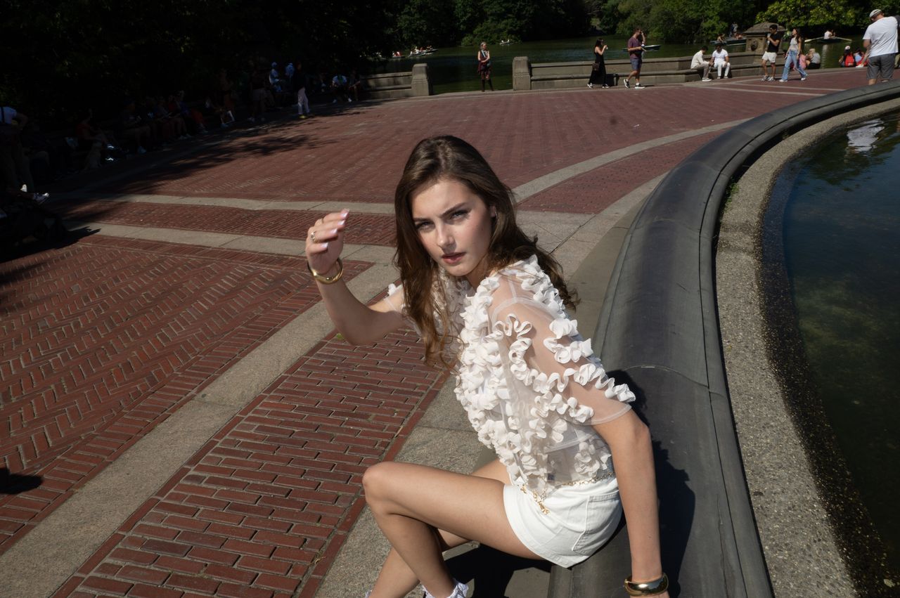 Mia Fantalin leaning against the stone rim of Bethesda Fountain in Central Park, in a sheer white ruffled top and white shorts